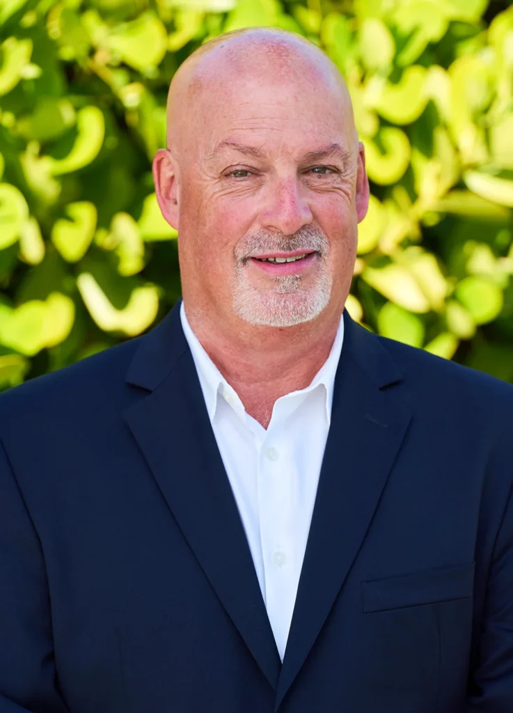Professional headshot of a smiling bald businessman with salt-and-pepper beard, dressed in navy suit and white shirt, against lush green tropical foliage background.