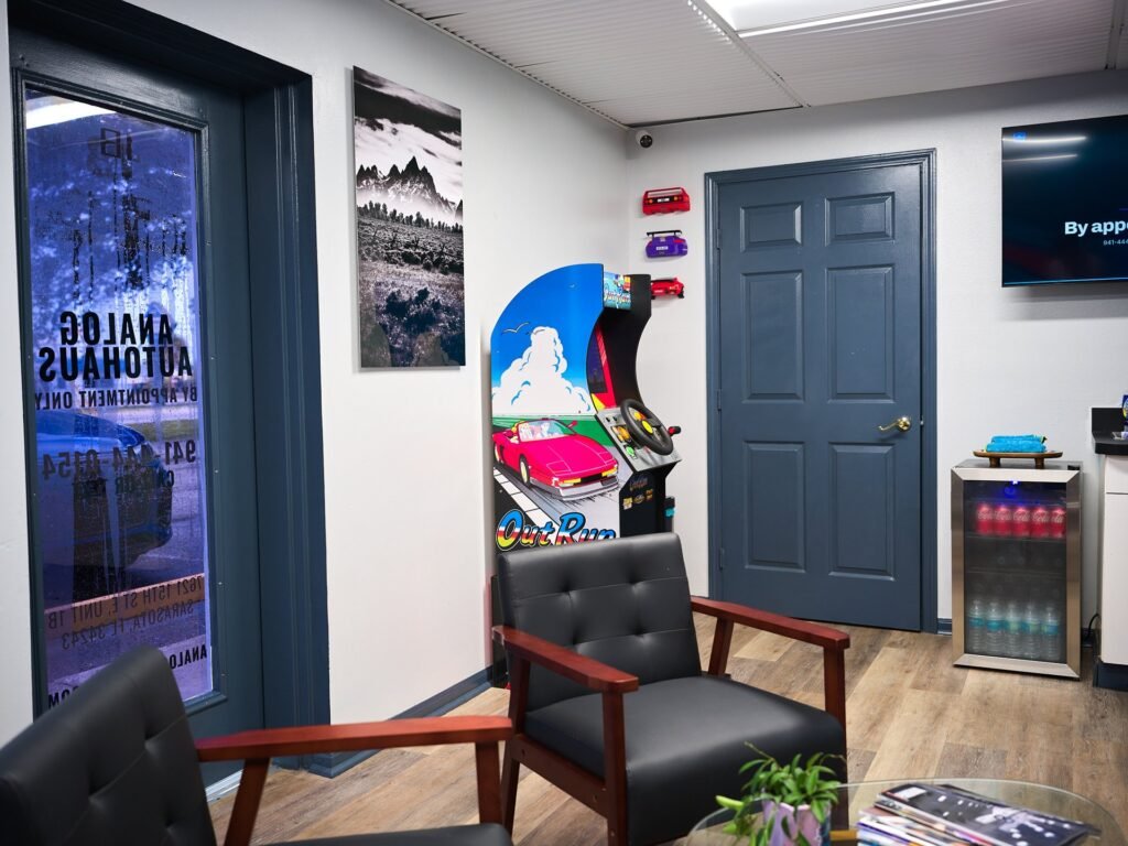 Analog Autohaus Sarasota office waiting area with OutRun arcade cabinet, fine-art mountain print by Nicholas, lettered glass door and leather armchairs. Photo by Thru My Eyes Productions.