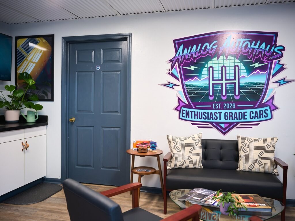 Analog Autohaus Sarasota lounge with retro neon Enthusiast Grade Cars logo mural above black leather mid-century loveseat. Photo by Thru My Eyes Productions.